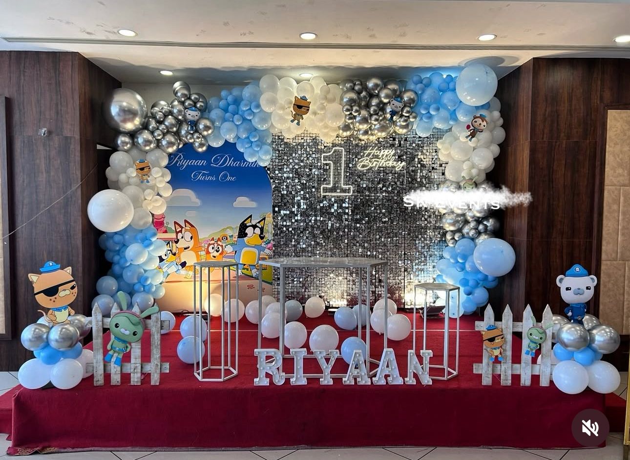 Cartoon-Themed 1st Birthday Party Decoration Kit with character cutouts, silver sequin backdrop, blue and white balloon garland, personalized name display, geometric pedestals, and white picket fences.