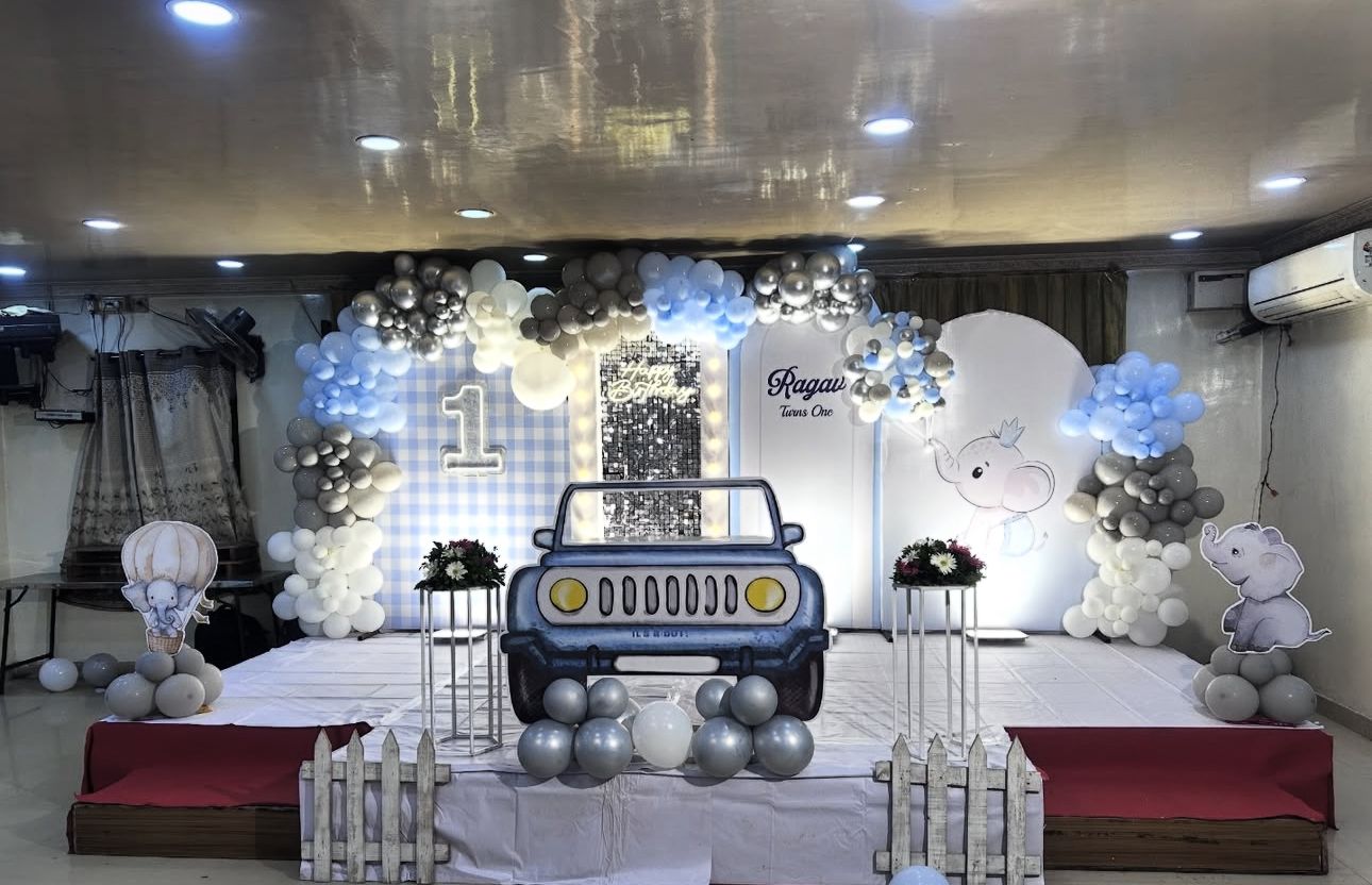 Elephant-Themed 1st Birthday Decoration Kit with pastel balloon arch in grey, blue, white, and silver, personalized name display “Ragav Turns One,” cartoon elephant cutouts, gingham backdrop, car cutout centerpiece, and LED Happy Birthday light sign.
