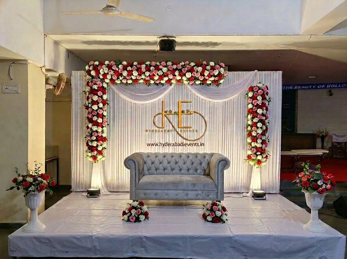 White floral stage decoration with elegant backdrop, red and pink flowers and modern sofa seating.