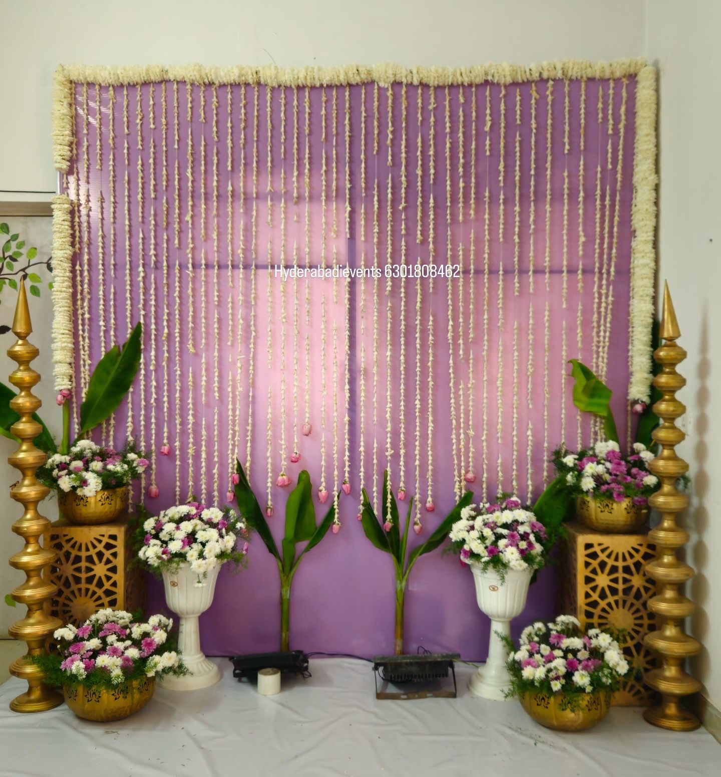 Minimal jasmine curtain backdrop decoration with purple background, brass lamps and floral arrangements for traditional events in Hyderabad.