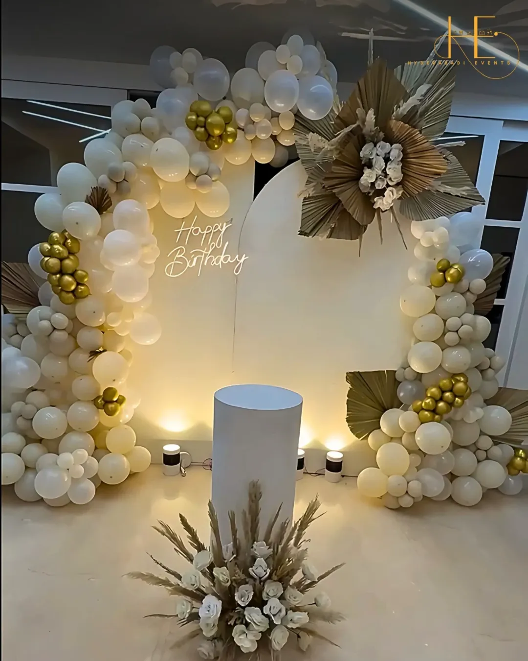 Elegant birthday decoration with white and gold balloons, floral elements and happy birthday neon sign in Hyderabad.