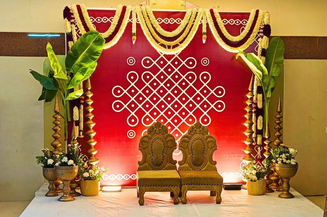 Traditional red Seemantham stage decoration with Muggu backdrop, banana leaves, brass lamps and carved wooden chairs in Hyderabad.