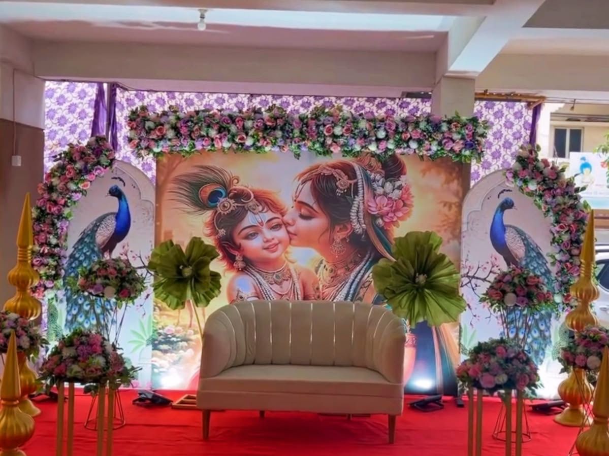 Premium Radha Krishna baby shower backdrop with floral arches, peacock panels, luxury sofa and elegant floral stage decoration.