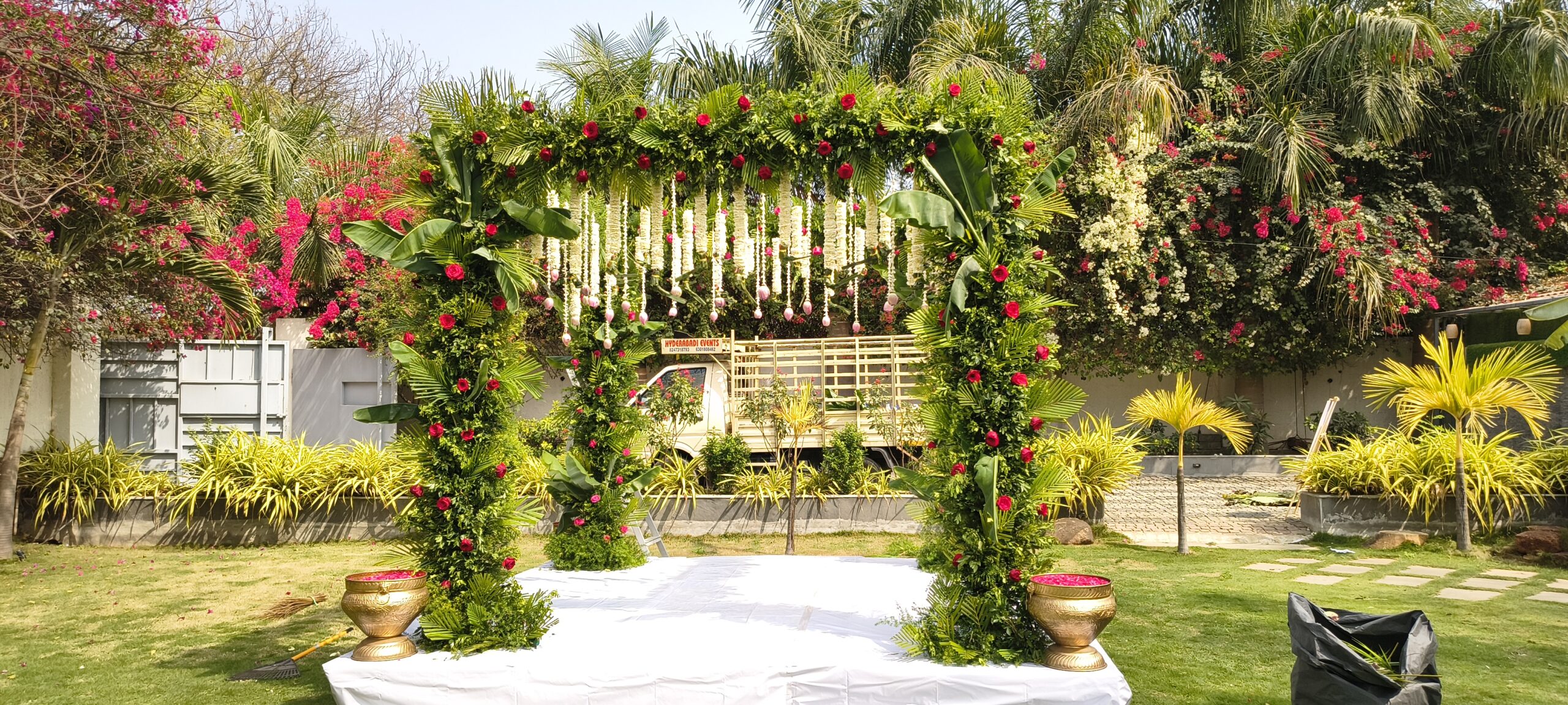 Luxury outdoor wedding entrance decoration with green foliage arch, red roses, banana leaves, and hanging jasmine garlands by Hyderabadi Events in Hyderabad.
