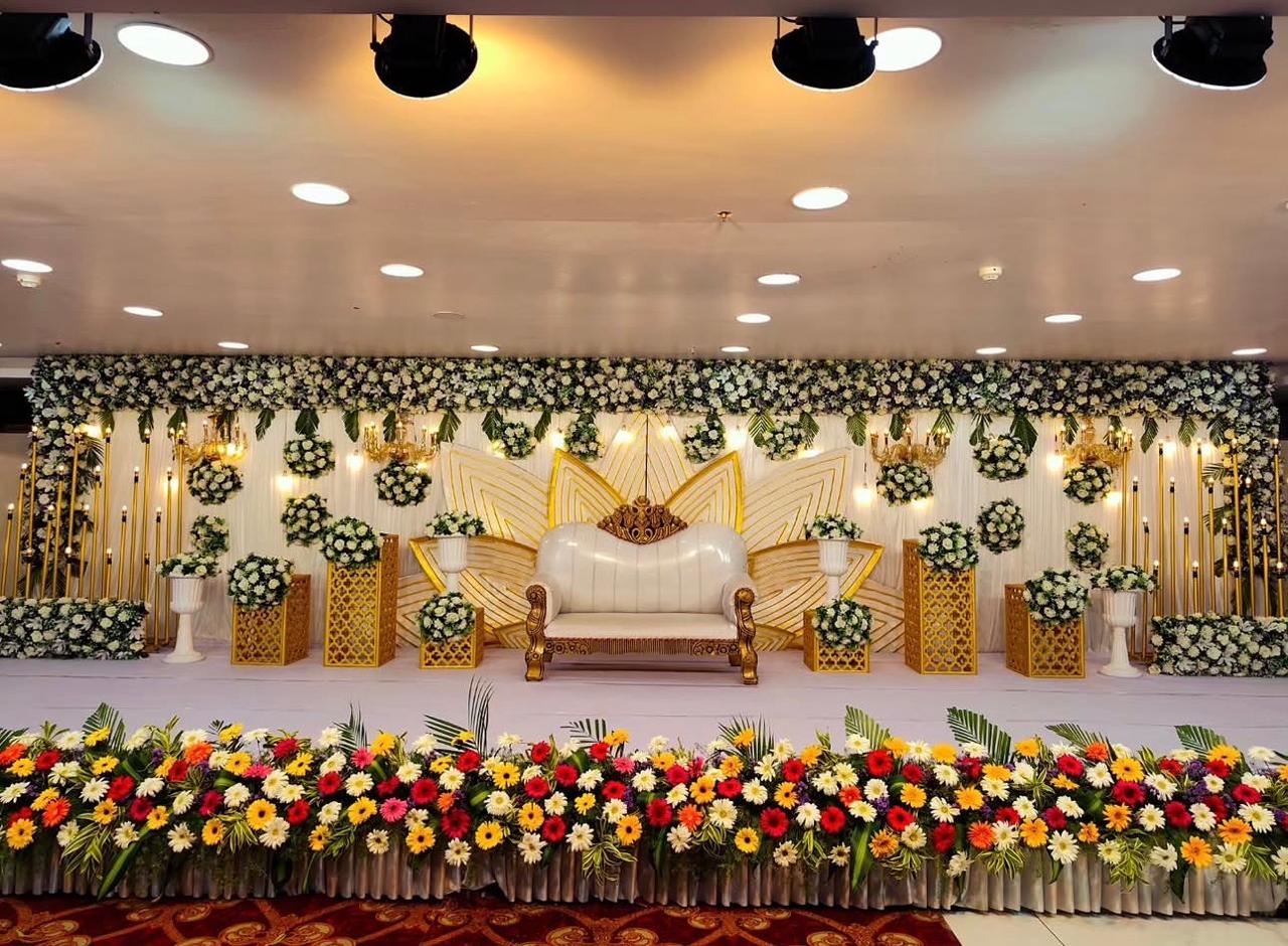 Grand white and gold wedding reception stage with a decorative lotus-inspired backdrop, ornate white sofa, hanging floral arrangements and colourful flower border.