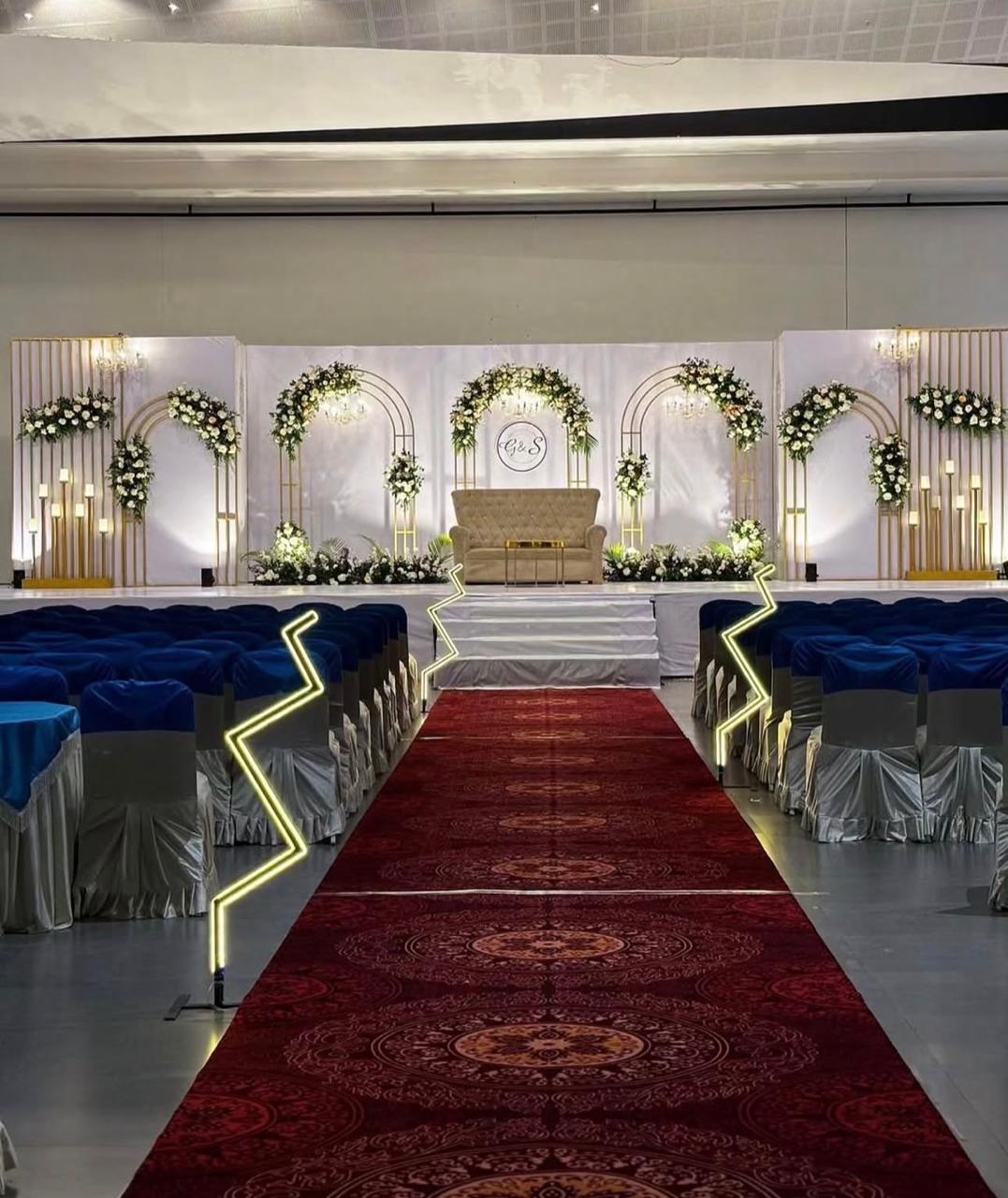 White and gold wedding stage with floral arches, a beige sofa, gold frame decorations, blue guest seating and a red carpet aisle.