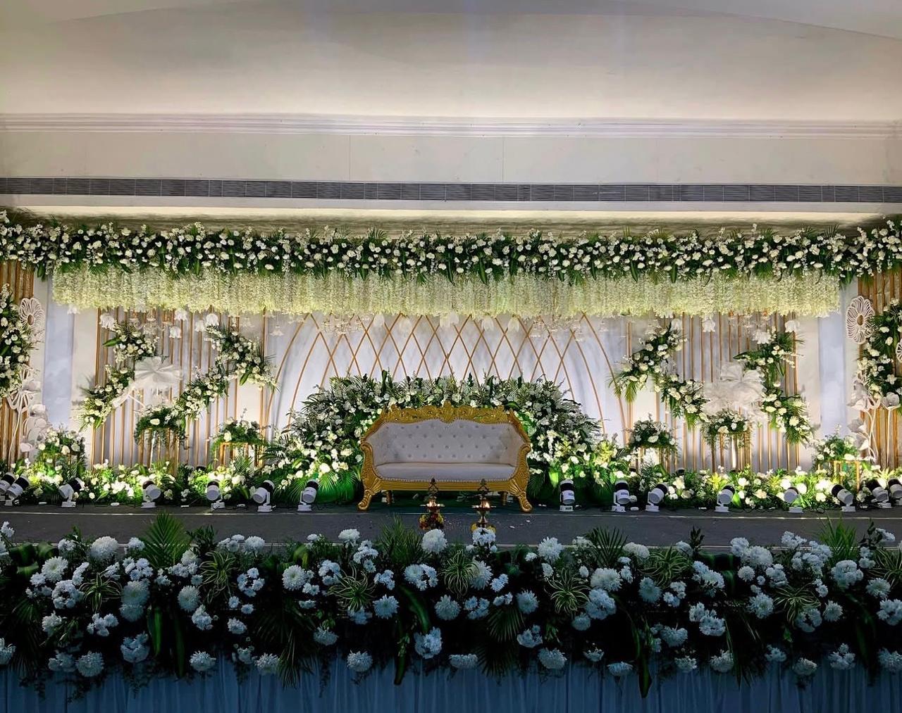 White and gold reception stage with an ornate central sofa, geometric gold backdrop, hanging white floral arrangements and decorative gold planters.