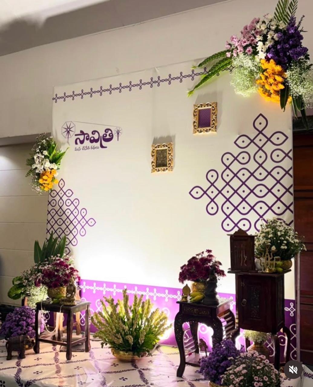 White Telugu traditional function backdrop with purple muggu patterns, framed decorative panels, colourful flower arrangements and wooden display stands.