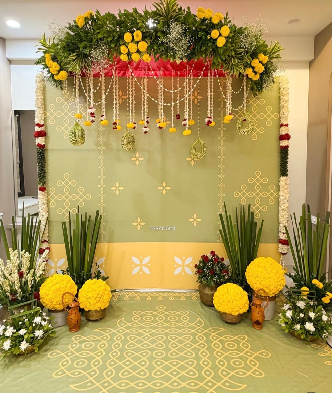 Green and yellow traditional pooja stage with a leafy floral canopy, hanging white flower strings, yellow marigold arrangements and a decorative kolam floor design.