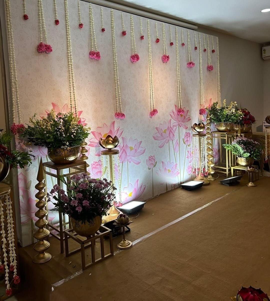 White botanical backdrop with hanging jasmine garlands and pink floral clusters, gold decorative planters and traditional floral arrangements.