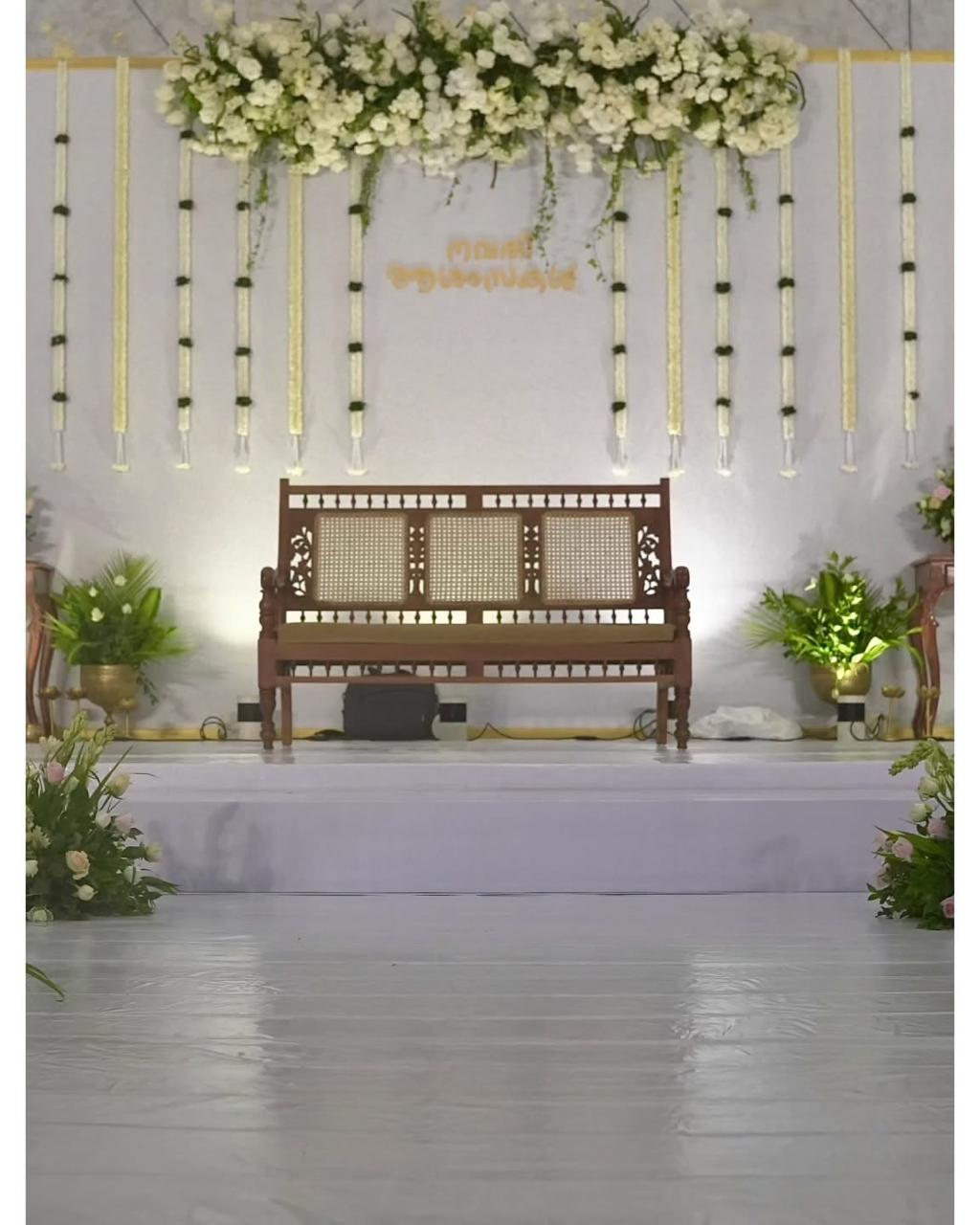 Traditional wooden sofa on a white event stage with a white flower canopy, hanging jasmine garlands and green potted plants.