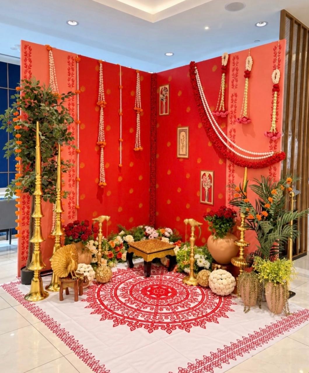 Traditional red and coral ceremony backdrop with hanging jasmine garlands, brass oil lamps, floral arrangements and decorative floor mandala.
