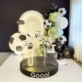 Soccer themed first birthday party decoration with neon number “1”, black and green balloon arch, large soccer ball props, and custom name backdrop for a festive celebration.