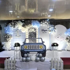 Elephant-Themed 1st Birthday Decoration Kit with pastel balloon arch in grey, blue, white, and silver, personalized name display “Ragav Turns One,” cartoon elephant cutouts, gingham backdrop, car cutout centerpiece, and LED Happy Birthday light sign.